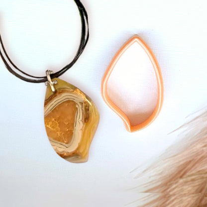 A teardrop-shaped pendant necklace made from a faux polished agate slice of polymer clay hanging on a black cord, shown next to its peach-colored PLA 3D printed clay cutter on a white background.
