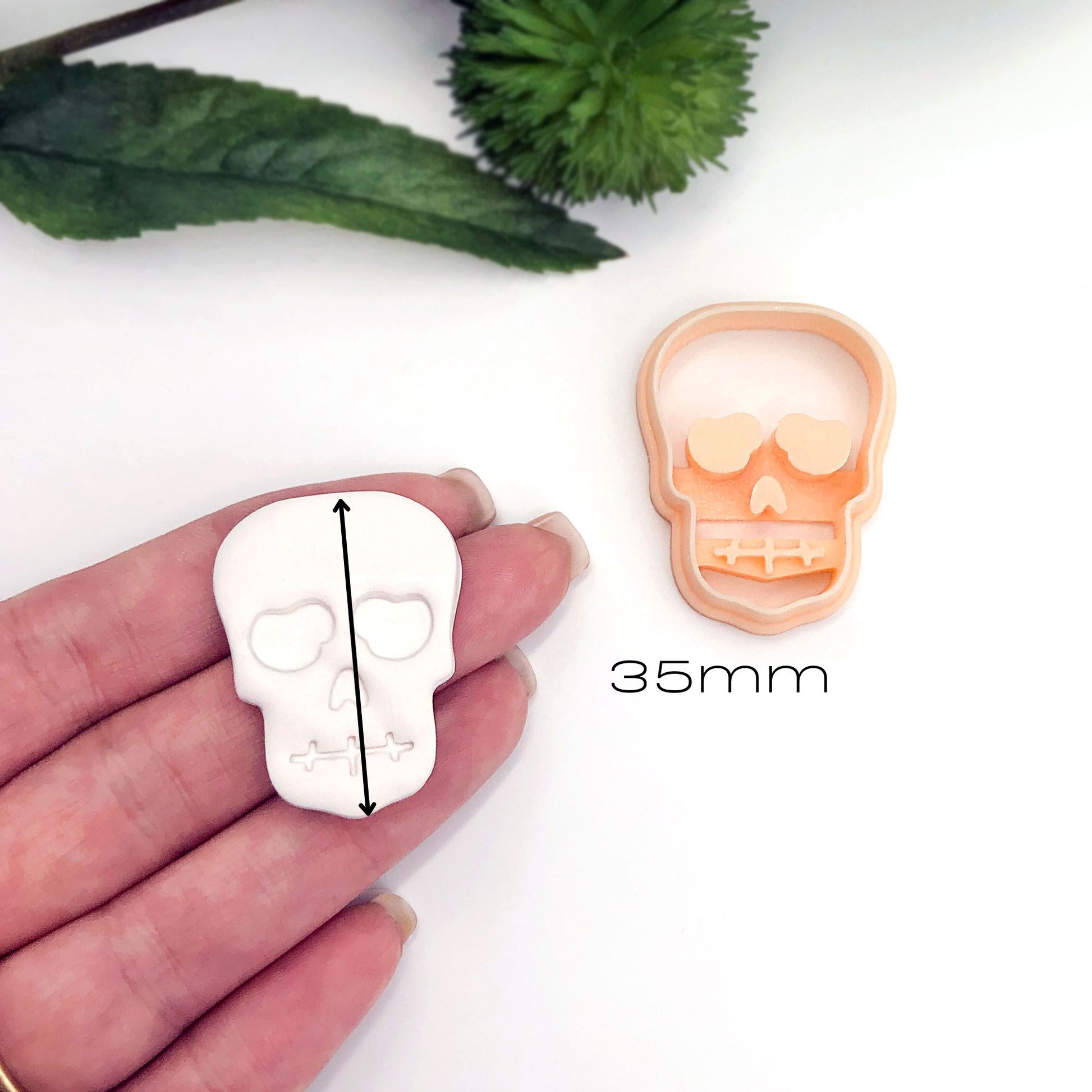 A hand holds a white skull-shaped item for scale next to a peach-colored skull-shaped mold or cutter, with a black measurement arrow and text reading 35mm on a white background.
