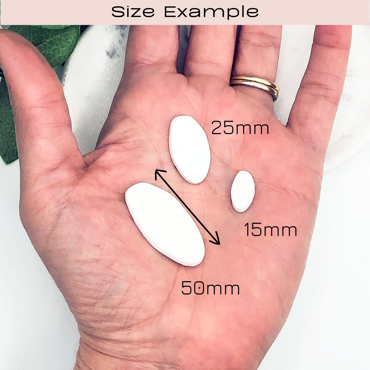 A hand displaying three white oval polymer clay cut outs of different sizes with black dimension labels reading 50mm, 25mm, and 15mm, under a header that says 'Size Example'.