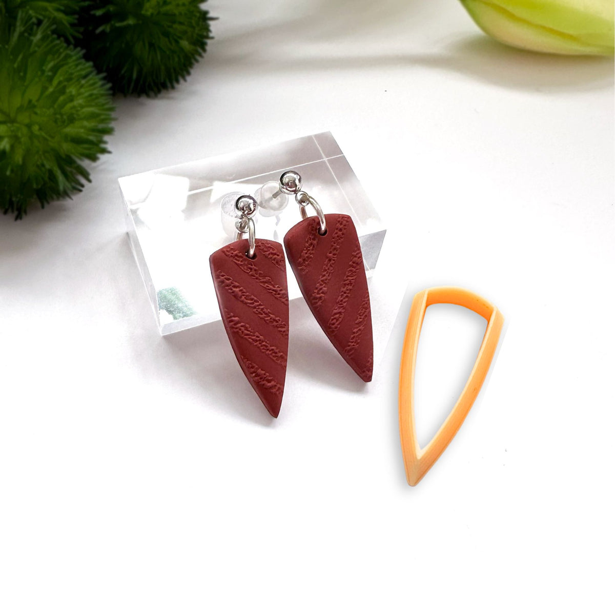 A pair of handmade dangle earrings with textured reddish-brown teardrop-shaped charms and silver posts, displayed on a clear acrylic stand next to an orange plastic clay cutter on a white background.