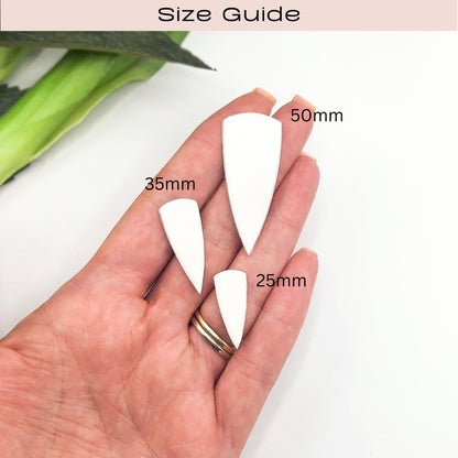 A hand holding three white pointed dagger drop clay cutouts labeled 50mm, 35mm, and 25mm under a 'Size Guide' header, with a soft-focus green leaf in the background.