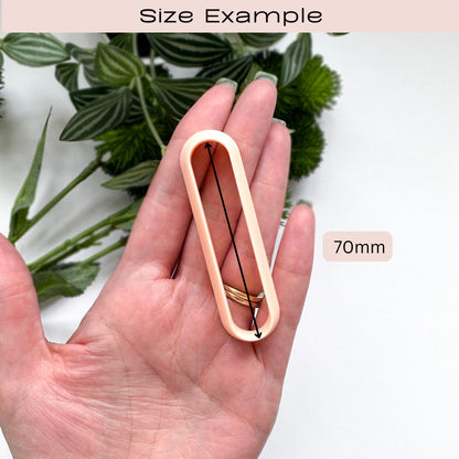 A hand holds a peach-colored clay cutter with a 70mm size indicator and text reading 'Size Example' and '70mm'. Green leaves are blurred in the background.