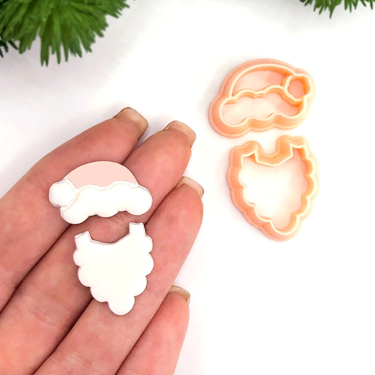 A hand holds two small Santa-themed cut outs, one in pale pink and one in peach, next to two matching cutters on a white background with small green foliage in the corner.