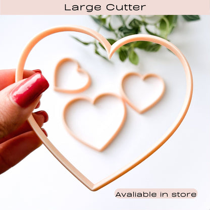 A hand with red nail polish holds a large peach-colored heart-shaped cookie cutter over a white background, with three smaller matching heart cutouts blurred behind it. Text at the top reads 'Large Cutter' and text at the bottom says 'Availiable in store'.
