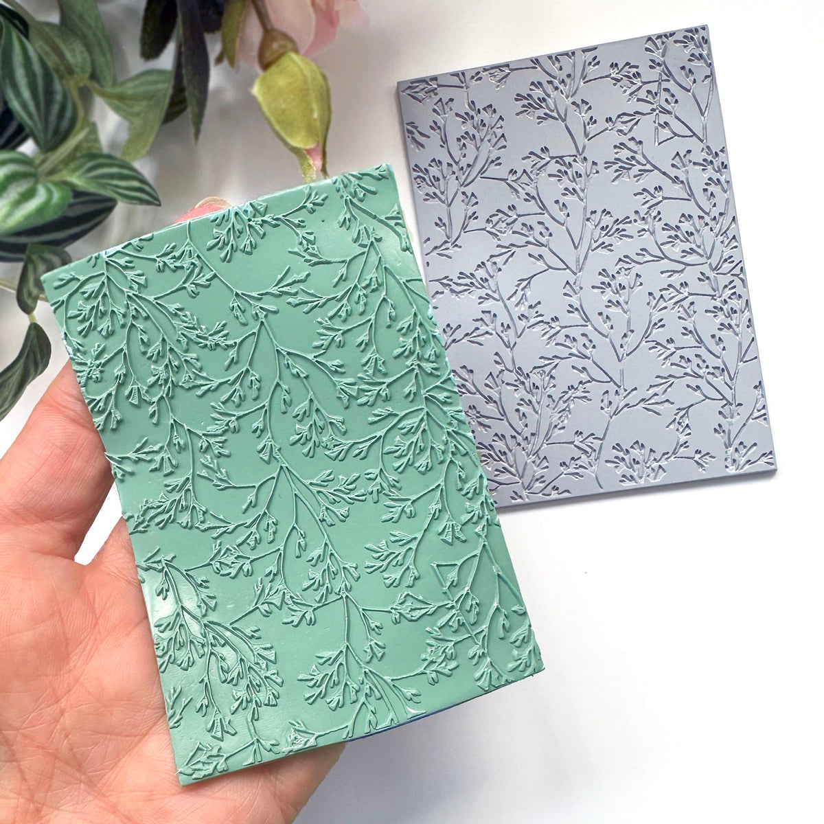 A hand holds a mint green polymer clay embossed surface made with the grey coral leaf stamp placed on a white surface in the background.