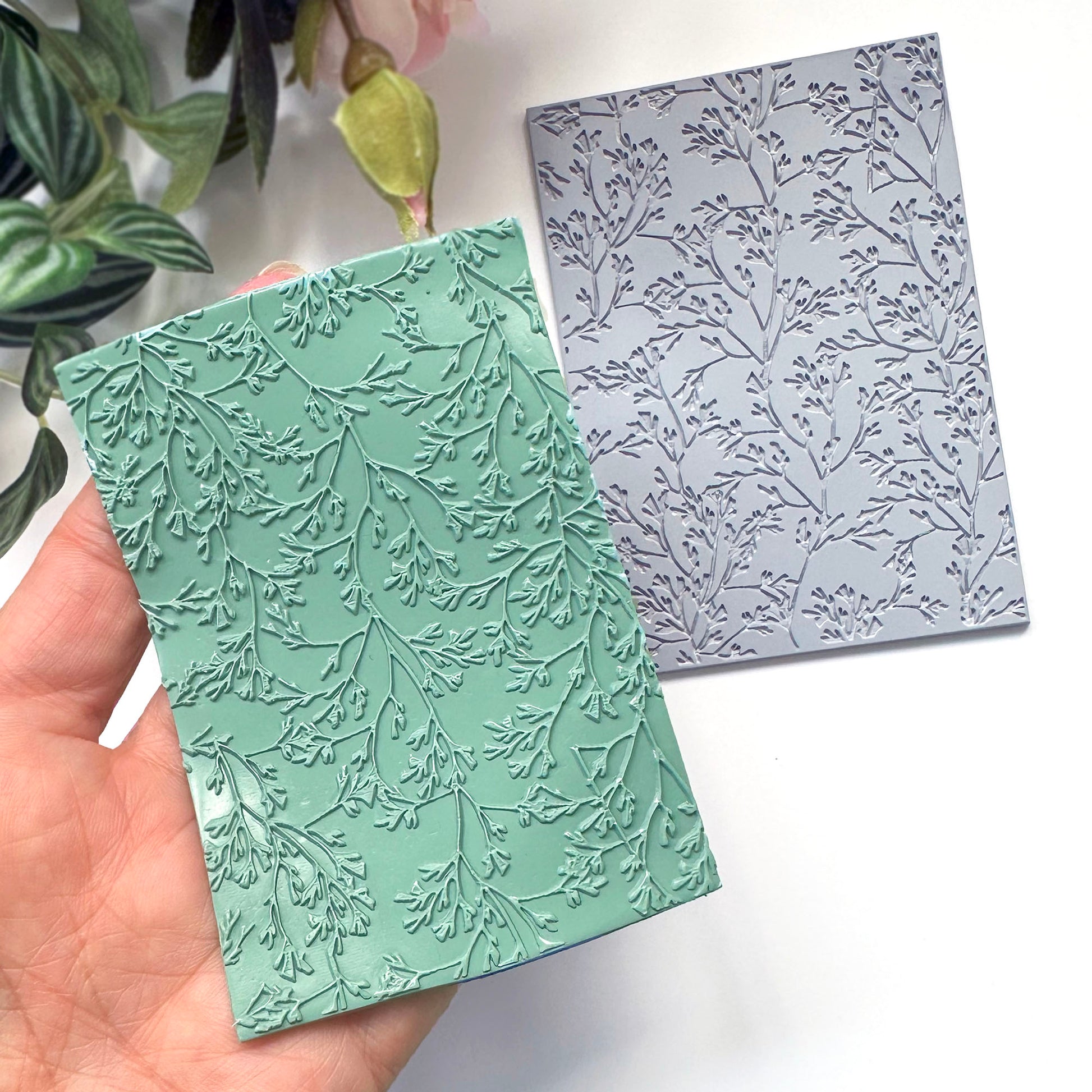 A hand holds a mint green polymer clay embossed surface made with the grey coral leaf stamp placed on a white surface in the background.