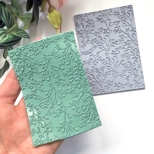 A hand holds a mint green polymer clay embossed surface made with the grey coral leaf stamp placed on a white surface in the background.