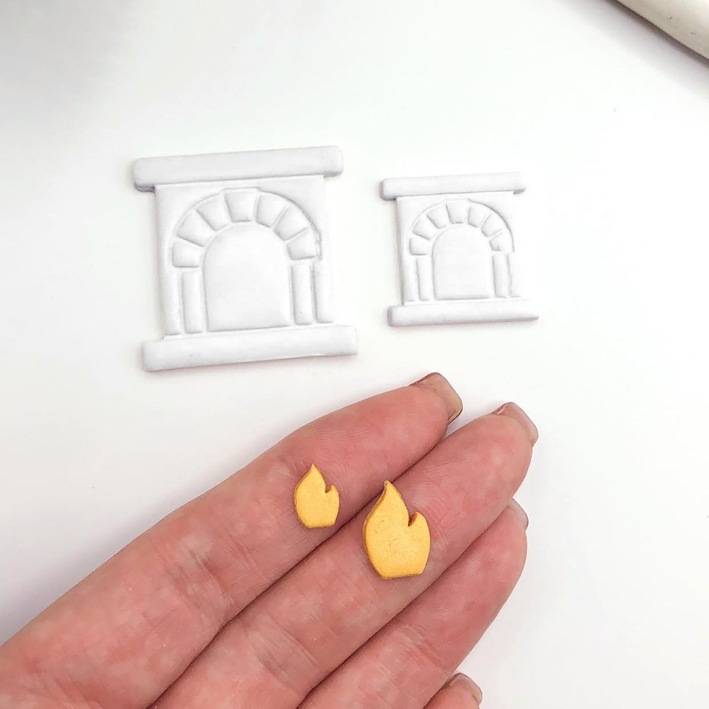 Two small yellow flame-shaped pieces held on a person's fingers, with two white fireplace-shaped polymer clay cut outs above on a plain white background.