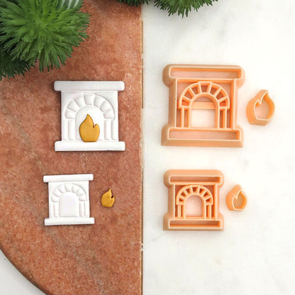 Two peach-colored fireplace cookie clay cutters and two white polymer clay cut outs each paired with a small gold flame, arranged on a split brown and white background with green pine branches in the corner.