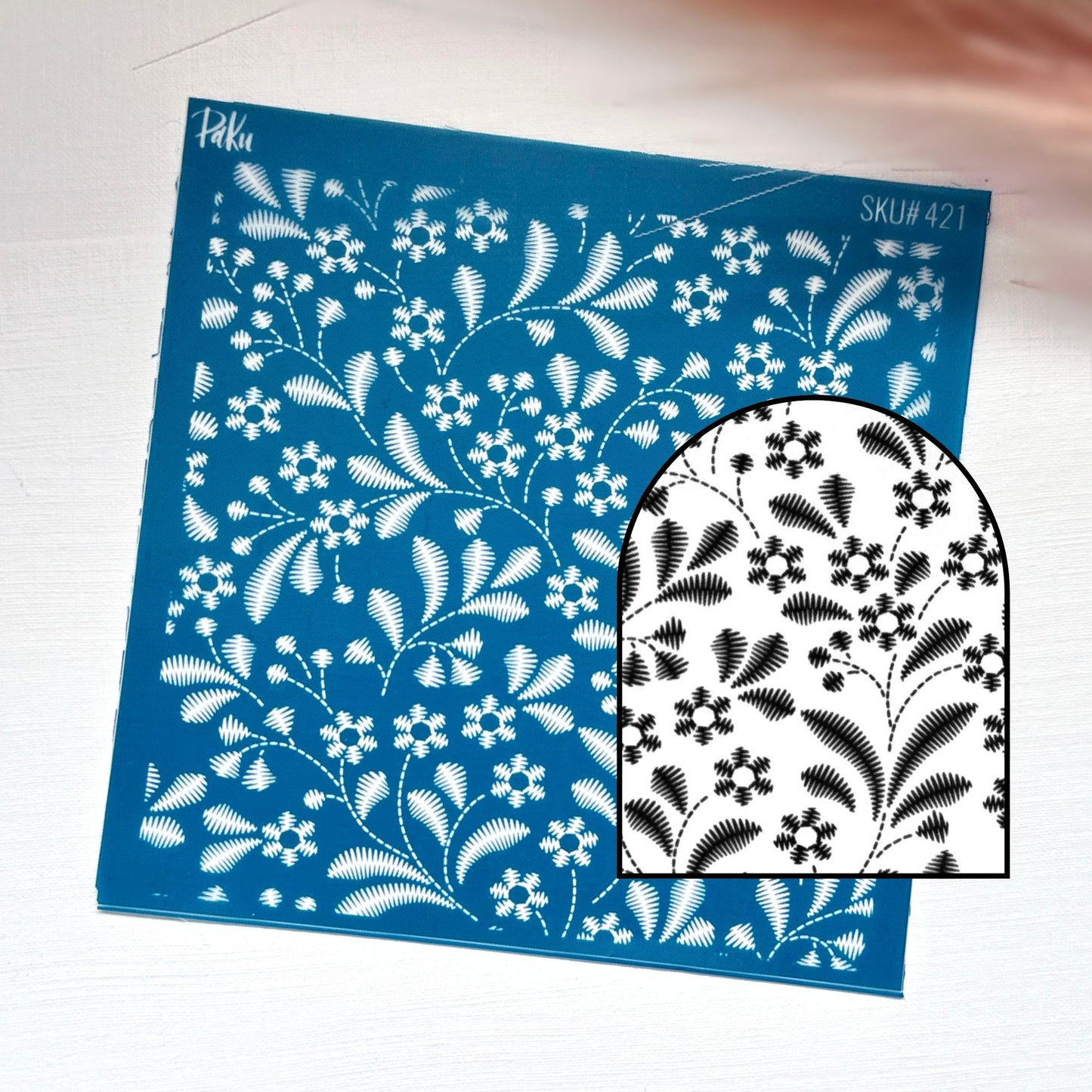 A blue rectangular silkscreen stencil sheet with a white floral and leaf pattern, shown on a light surface with a smaller inset showing the black-and-white design through the stencil.