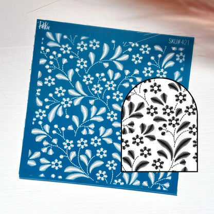 A blue rectangular silkscreen stencil sheet with a white floral and leaf pattern, shown on a light surface with a smaller inset showing the black-and-white design through the stencil.