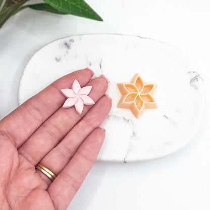 A hand holds a small pale pink poinsettia flower-shaped clay cutout next to a peach-colored flower-shaped cookie cutter on a white marble tray.