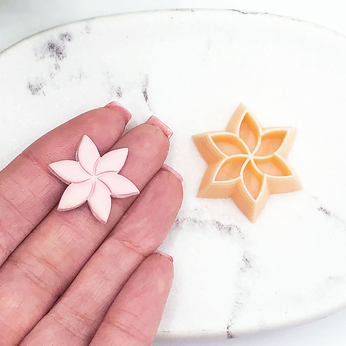 A small pale pink flower-shaped clay cut out resting on a person's fingers next to a peach-colored matching flower-shaped cutter on a white marble surface.