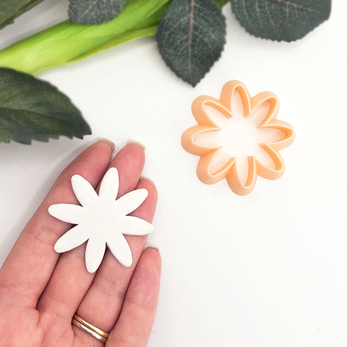 A hand holds a white clay flower cutter, with an orange plastic flower-shaped cookie cutter placed nearby on a white surface, with green leaves in the background.