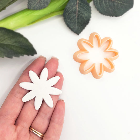 A hand holds a white clay flower cutter, with an orange plastic flower-shaped cookie cutter placed nearby on a white surface, with green leaves in the background.