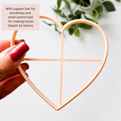 A hand with red-painted nails holds a peach-colored heart-shaped clay cutter against a white background with a soft-focus plant in the corner. 
