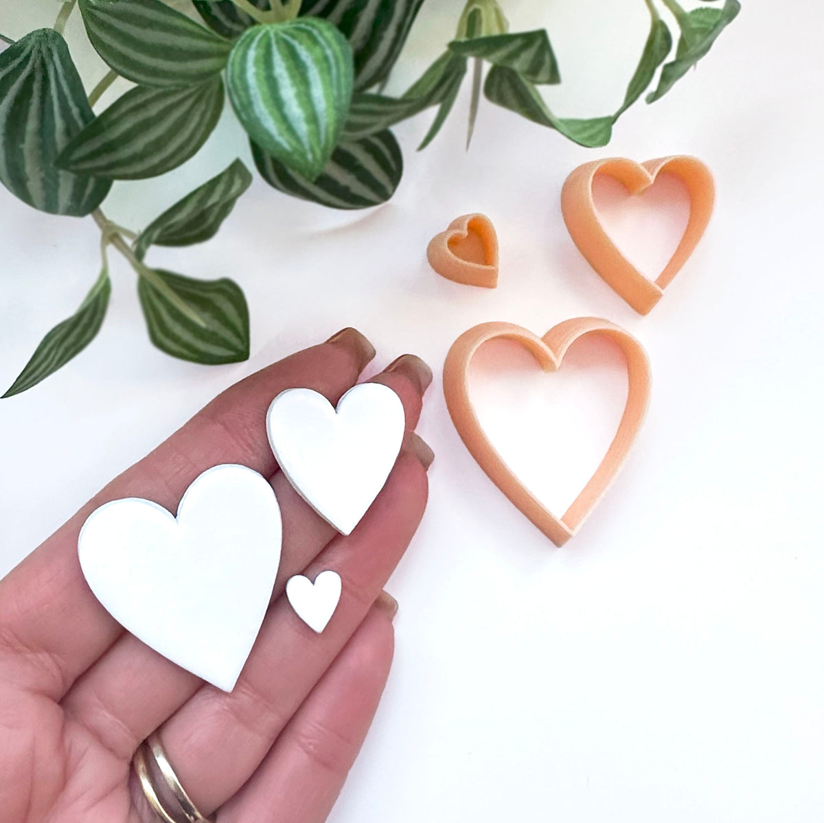 Heart-shaped cookie clay cutters in peach-color, shown with a hand holding three white cutouts and green plant leaves in the background.