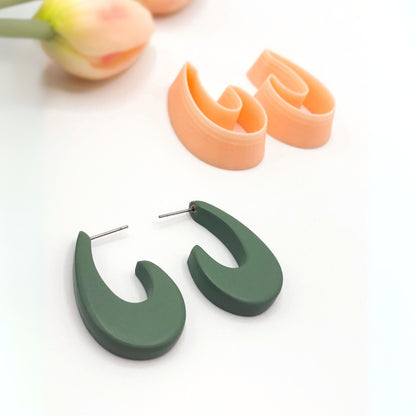 A pair of matte sage green organic droop-shaped hoop earrings with silver posts, displayed on a clean white surface next to a pair of peach-colored clay cutters and a blurred tulip in the background.