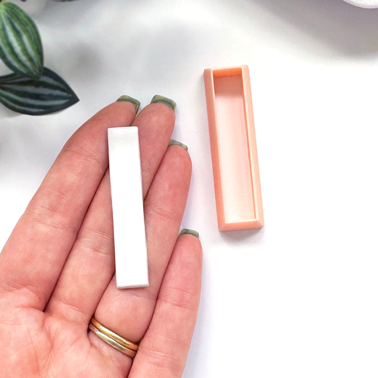 A hand holds a small white rectangular Polymer clay bar, with a peach-colored rectangular clay cutter beside it on a white surface.