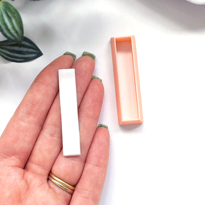 A hand holds a small white rectangular Polymer clay bar, with a peach-colored rectangular clay cutter beside it on a white surface.