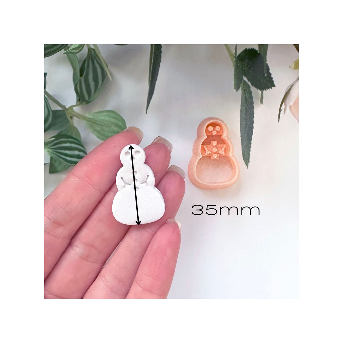 A hand holds a small white snowman-shaped clay cutout next to a peach-colored snowman cutter, with a black 35mm size marker shown on the white item. Green leaves are in the background.