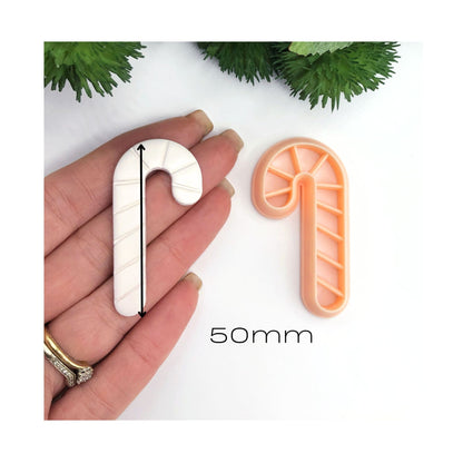 A candy cane-shaped cookie cutter shown next to a hand holding a matching white clay piece cutout for scale, with a black measurement arrow and the text '50mm' on a white background with green decorative foliage at the top.