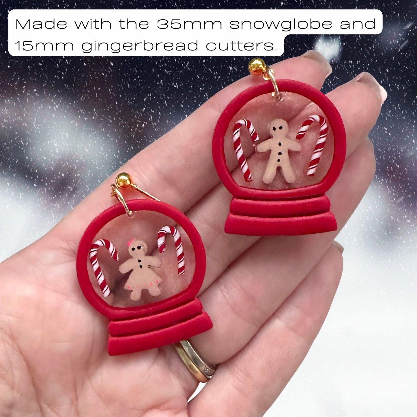 A pair of handmade Christmas snow globe earrings held in a person's hand. Each earring has a red circular frame with a small gingerbread figure and candy canes inside, suspended by gold rings.