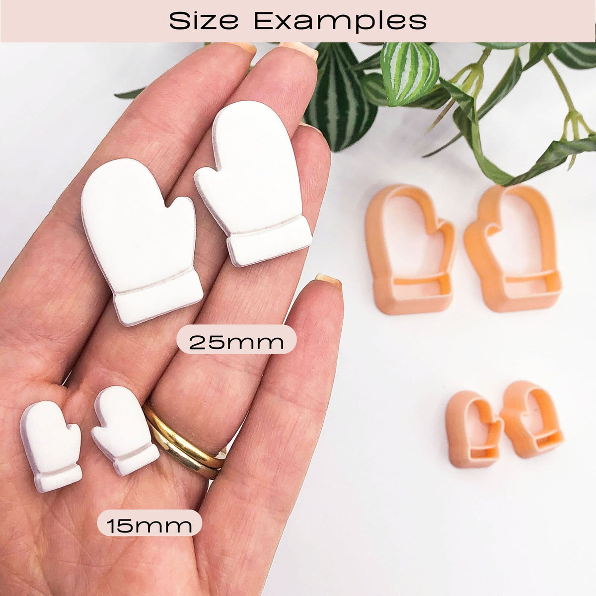 A hand holding 2 mirror sets of small white mitten-shaped clay cut outs, with two peach-colored mitten-shaped cookie cutters placed on a white surface nearby. A green plant leafy branch is visible in the upper right corner. With labels indicating 25mm and 15mm sizes under the heading 'Size Examples'.