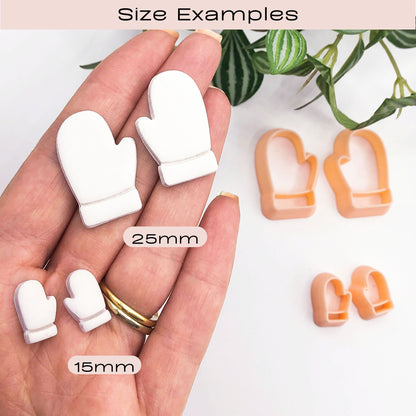 A hand holding 2 mirror sets of small white mitten-shaped clay cut outs, with two peach-colored mitten-shaped cookie cutters placed on a white surface nearby. A green plant leafy branch is visible in the upper right corner. With labels indicating 25mm and 15mm sizes under the heading 'Size Examples'.