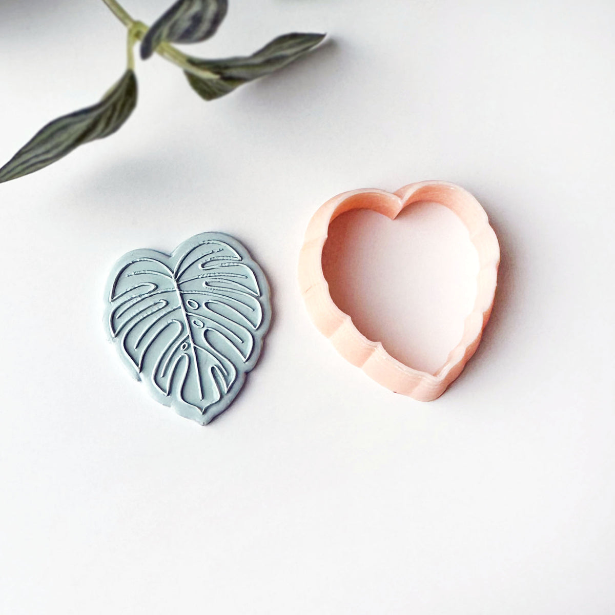 A monstera clay cutter in pale peach and a matching leaf embossed stamp in dusty blue-gray, displayed on a clean white surface with a decorative leaf sprig in the corner.
