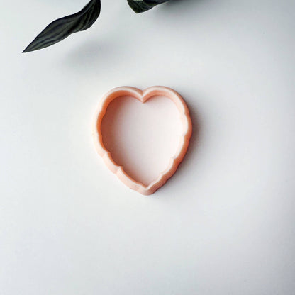 A monstera cookie cutter in a soft peach-pink color, shown on a clean white background with a decorative dark green leaf in the top-left corner.