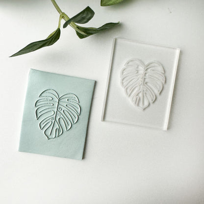 A monstera leaf embossed acrylic stamp shown stamped into blue clay, displayed on a clean white surface with a green plant stem in the background.