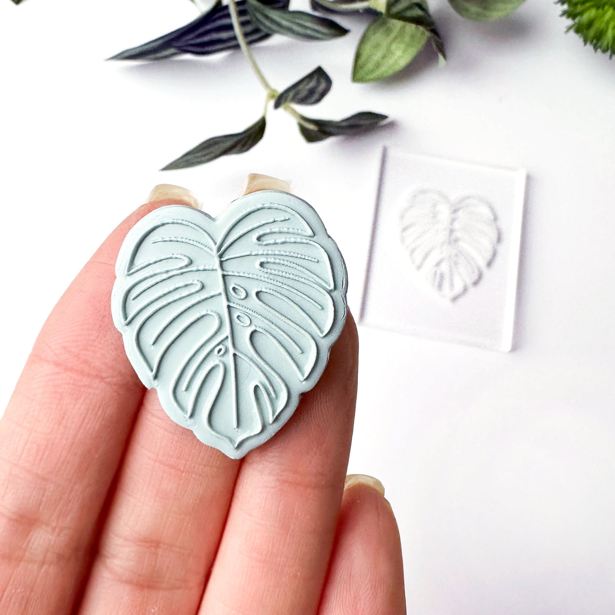 A monstera leaf stamped in clay held in a person's hand, with a clear acrylic block showing the stamped impression in the background on a white surface with green leaves.