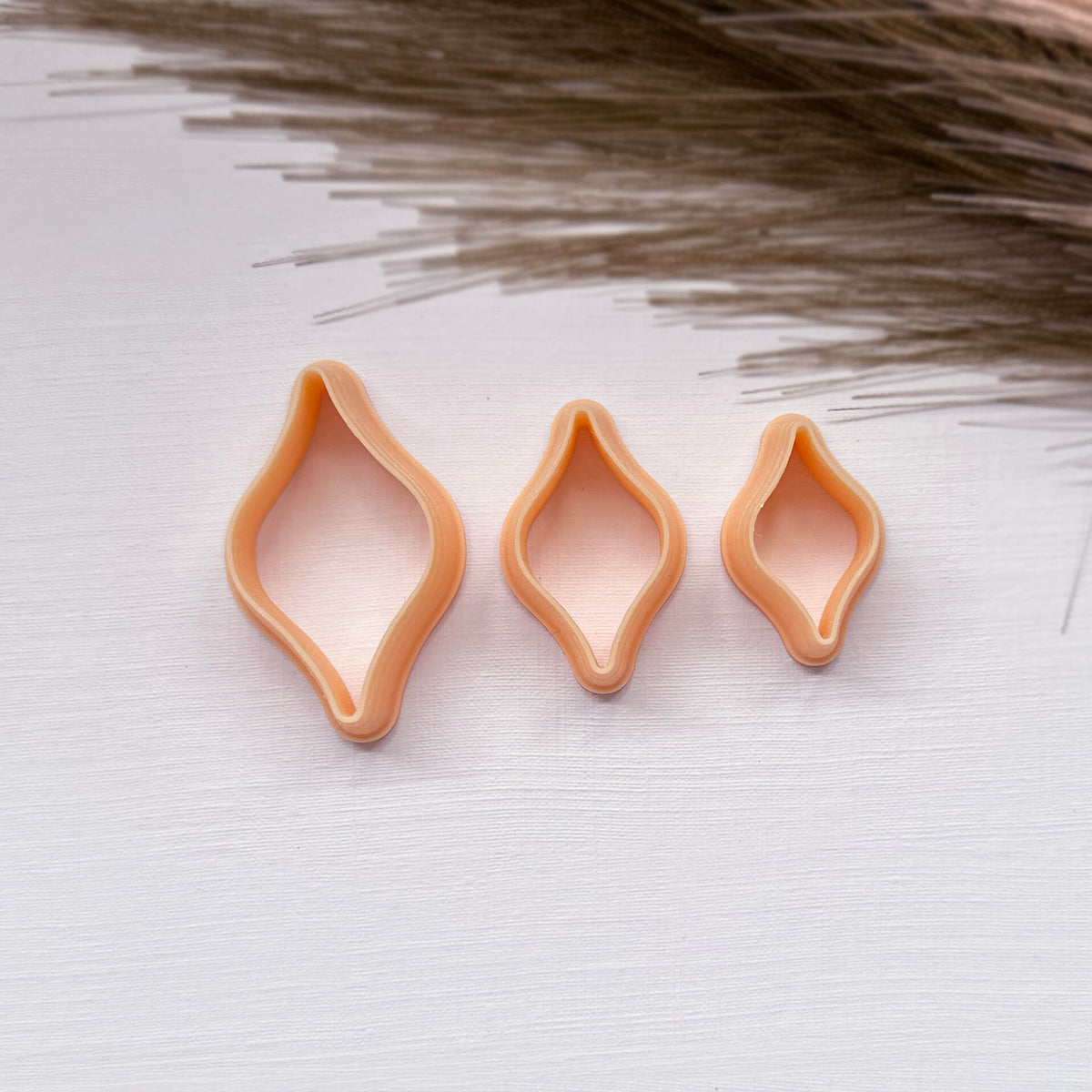 Three peach-colored ogee bulb clay cookie cutters in different sizes arranged on a white textured surface with dried grass in the background.