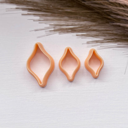 Three peach-colored ogee bulb clay cookie cutters in different sizes arranged on a white textured surface with dried grass in the background.