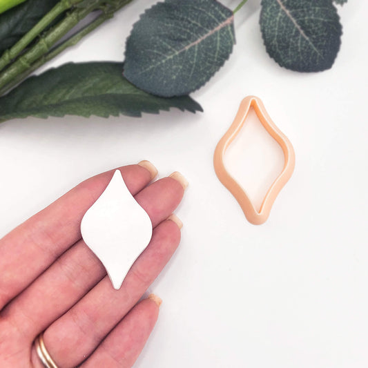 A hand holds a small white ogee bulb-shaped clay piece next to a peach-colored diamond-shaped cookie cutter on a white surface with green leaves in the background.
