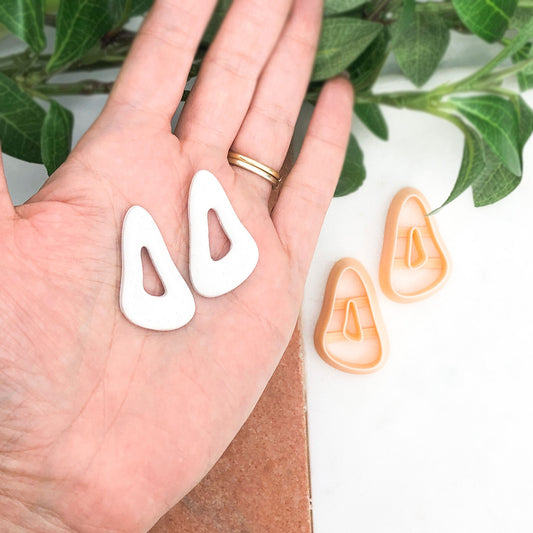 A hand holds two white organic pebble drop-shaped earrings, with two peach-colored plastic clay cutters placed on a white surface nearby. Green leaves and a brown textured mat are visible in the background.