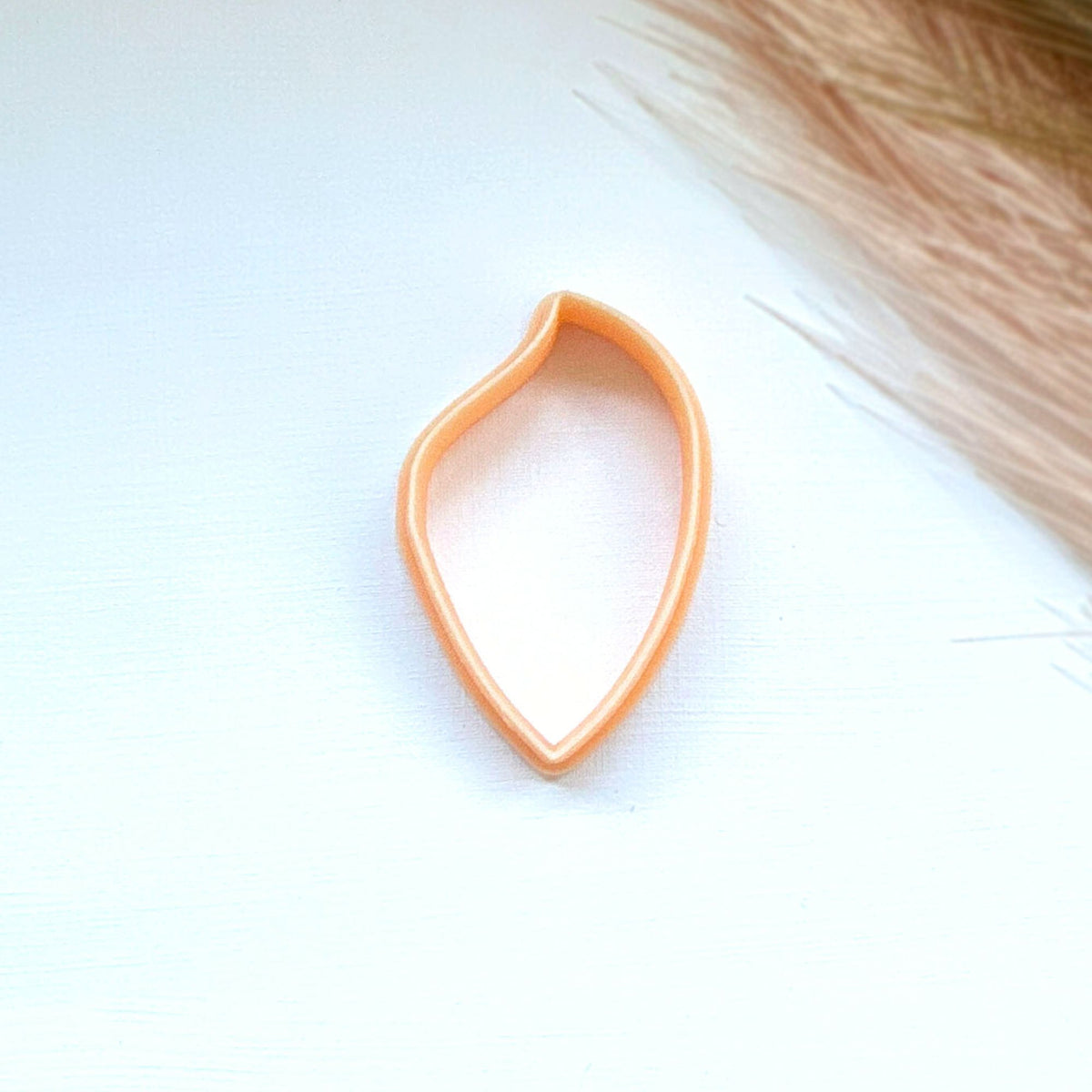 A small peach-colored clay cutter or cookie cutter shaped like a teardrop or leaf, shown on a white textured surface 