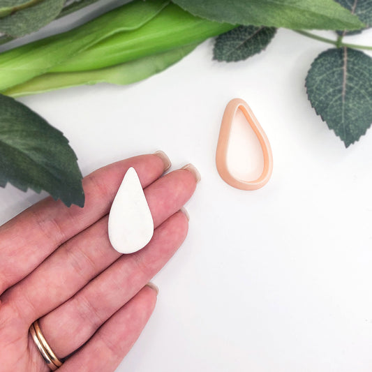 A hand holds a small white organic teardrop-shaped clay piece while another matching teardrop clay cutter in peach sits nearby on a white surface with green leaves in the background.