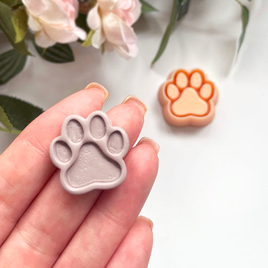A hand holds a small paw-shaped polymer clay cut out in a light taupe grey colour, with a paw-shaped clay cutter in peach in the background. Pink flowers and green leaves are softly blurred at the top.