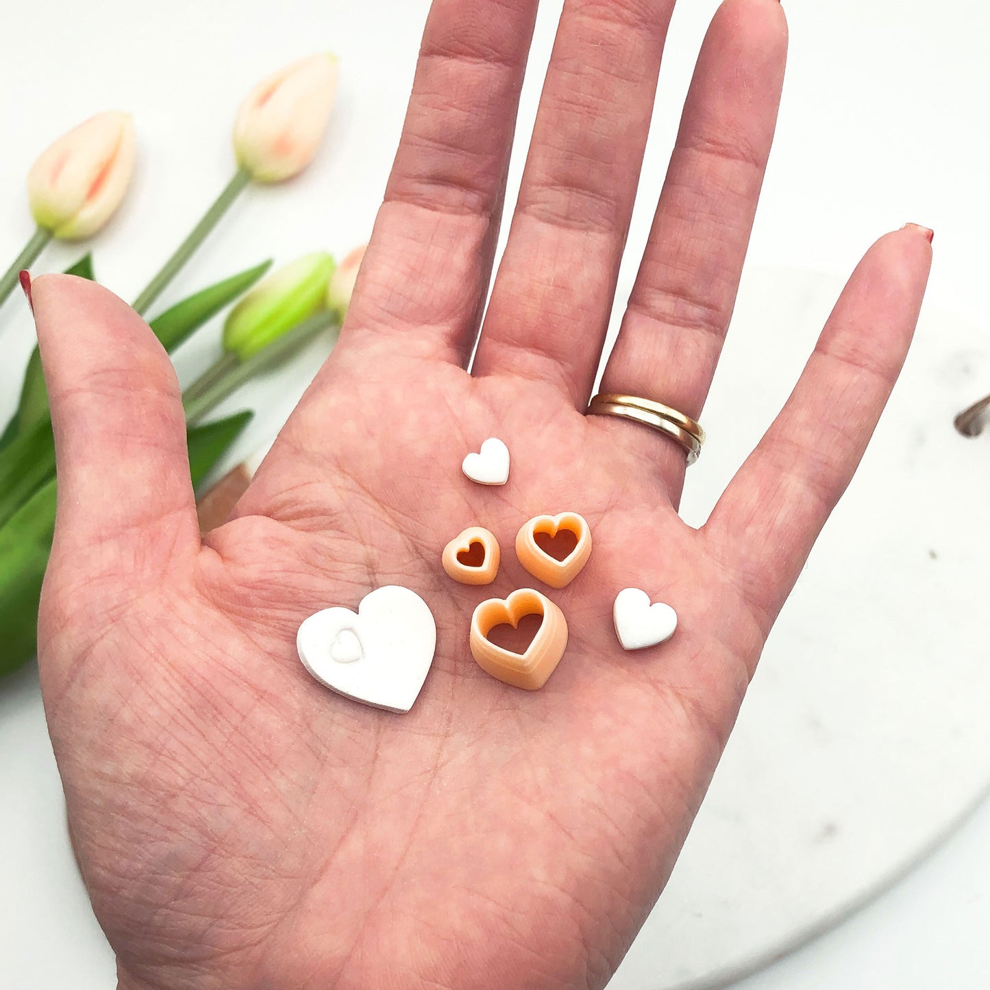 A hand holds several small heart-shaped jewelry pieces in white and peach-orange, with mini clay cutters with tulips blurred in the background.