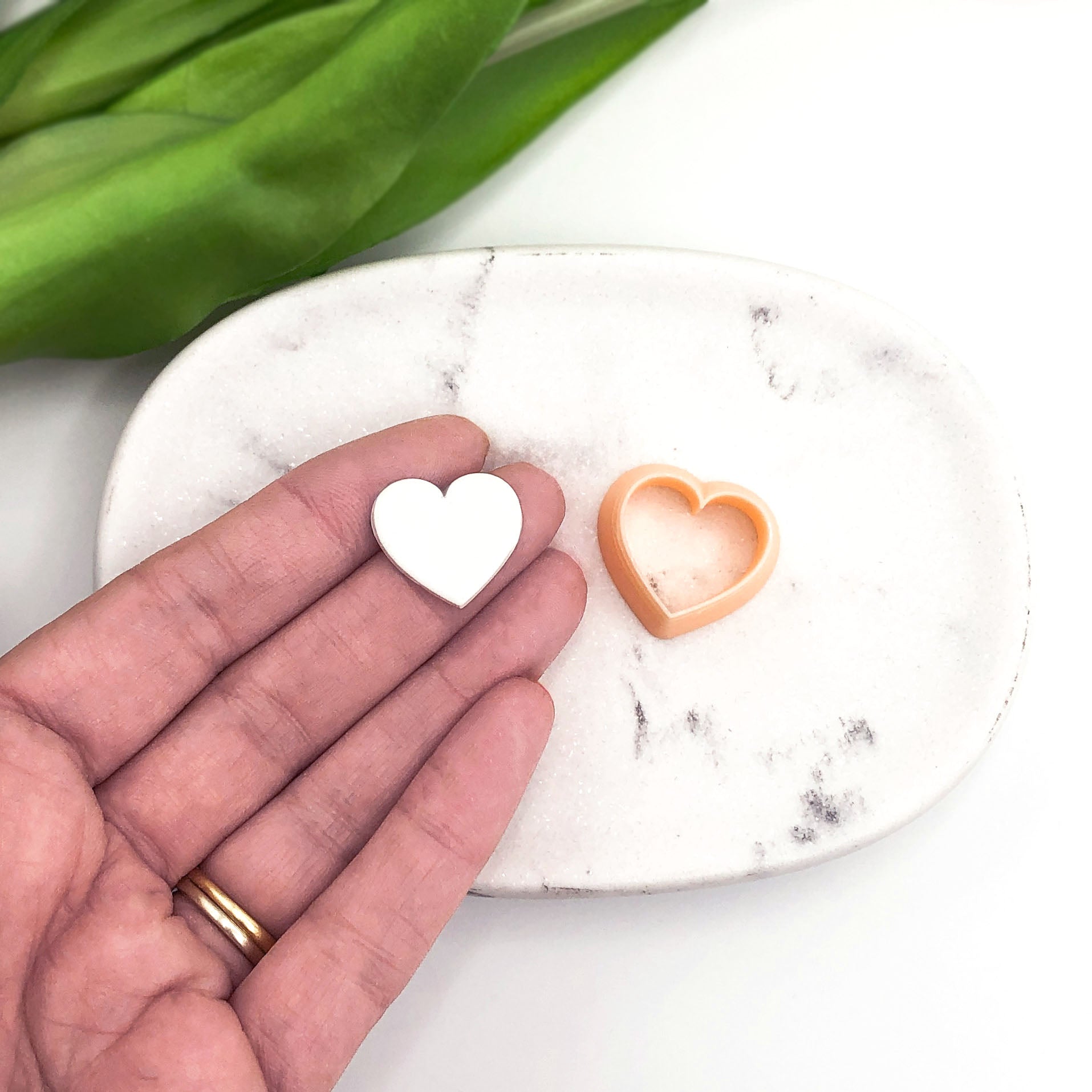 A hand holds a small white heart-shaped polymer clay cut out next to a peach-colored heart-shaped cookie cutter on a white marble tray, with green tulip leaves in the background.