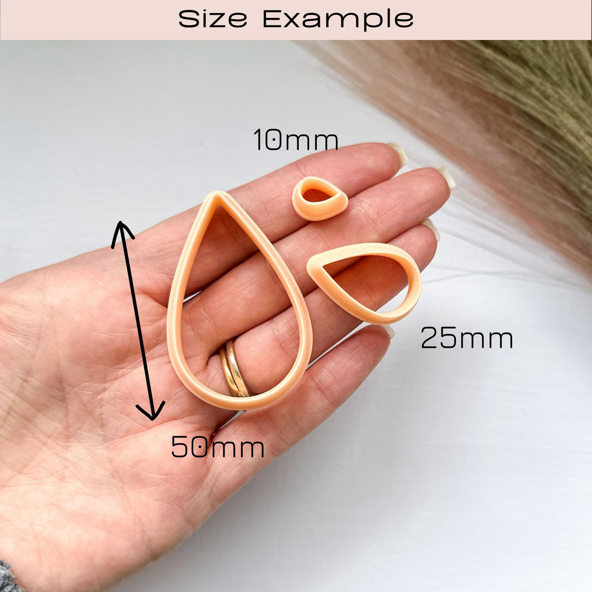 Size example of 3 peach-coloured plastic clay cutters sat on a hand in showing sizes   10mm, 25mm  and 50mm. The cutters are shaped like raindrops