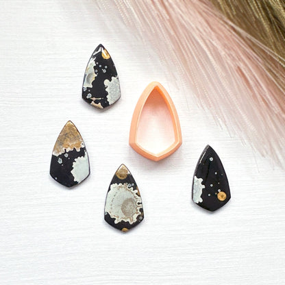Four pointed shield-shaped clay gemstones with black, white, and gold agate-like patterns are arranged around a peach-colored teardrop-shaped clay cutter on a white textured background with soft pink grass in the corner.