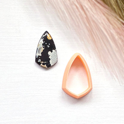A shield teardrop-shaped clay cut out  in black and white, and a peach clay cutter set against a light textured background with soft pink feather-like accents.
