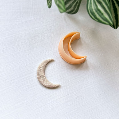 A crescent moon-shaped clay charm. Next to a peach-colored crescent moon clay cutter on a white textured background with green leaves in the corner.