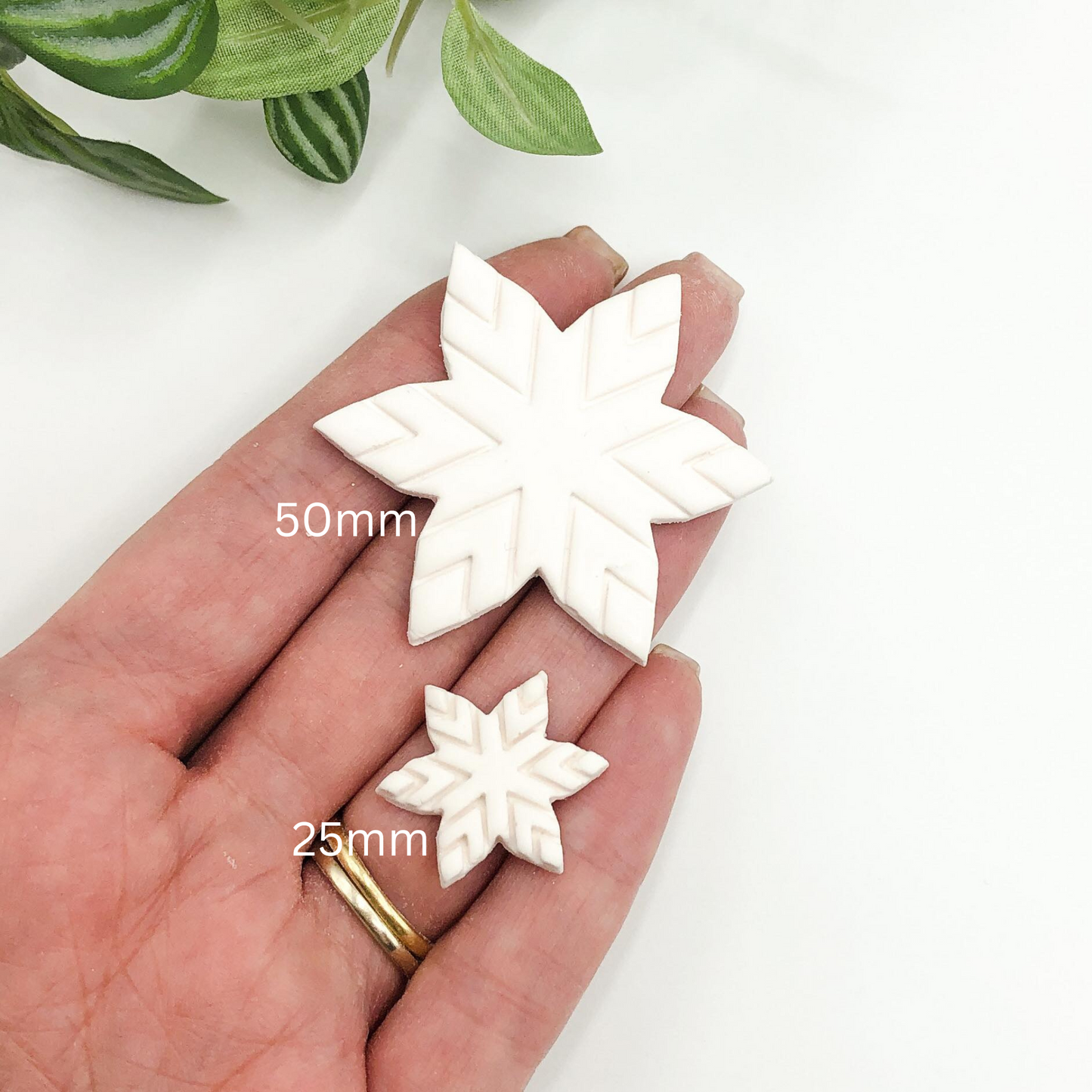 Two white snowflake-shaped polymer clay cutouts held in a person's hand, with size labels reading 50mm and 25mm. The hand is shown against a clean white background with a small green plant leaf in the corner.