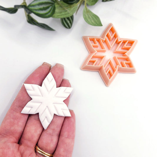 A hand holds a white snowflake-shaped polymer clay cut out while a peach-colored star-shaped cookie clay cutter sits on a white surface with green leaves in the background.