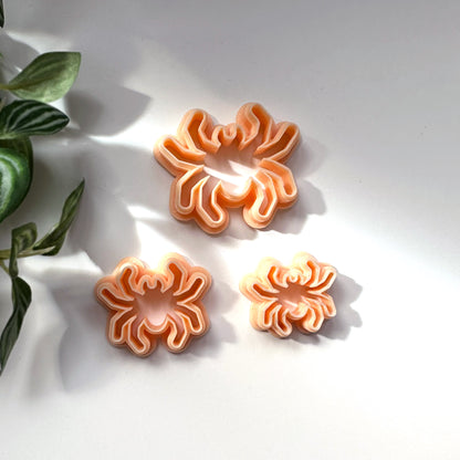 Three peach-colored plastic clay cookie cutters shaped like spiders, arranged on a white surface with a green plant leaf visible on the left.