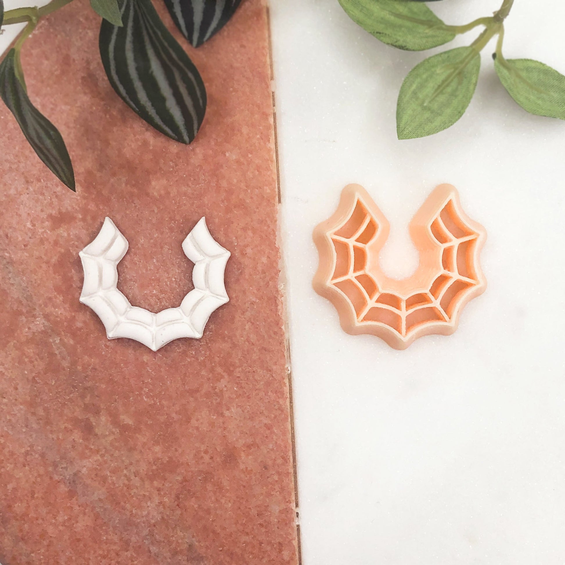 A small decorative spiderweb-shaped cookie clay cutter shown with one white polymer clay cut out on a  background with green leaves at the top.